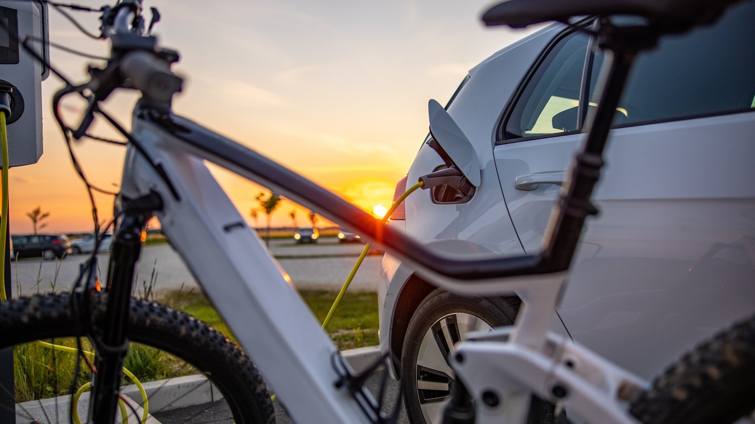 Sinnbild für Mobilität: Ein E-Auto vor einer Ladesäule, davor ein E-Bike, im Hintergrund ein Sonnenuntergang.