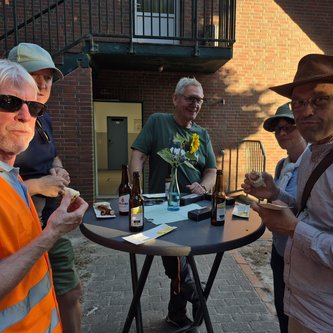 Fünf Tourteilnehmer*innen stehen um einen Tisch und schauen in die Kamera, einige davon mit Grillwürstchen in der Hand. Die Szene ist vor einem Gebäude am Hollener Badesee.